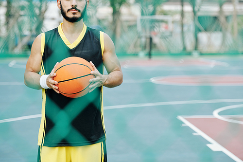 Young basketball player holding ball and looking through wire mesh fence around outdoor court