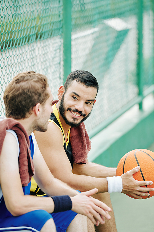 Smiling handsome young Hispanic man with basketball ball looking at friend telling funny story