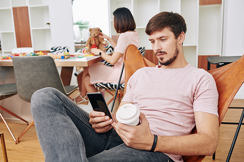 Handsome young man drinking coffee and texting friends when his wife is playing with daughter