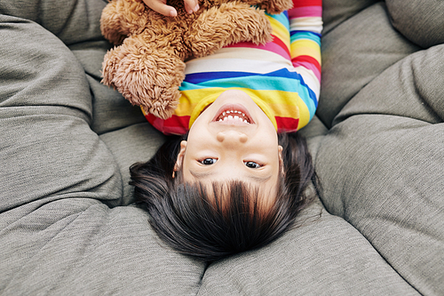 Excited happy young mixed race girl lying on duvet with teddy bear and looking at camera