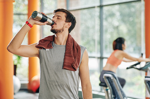 Thirsty sportsman with towel on shoulder drinking water after training in gym