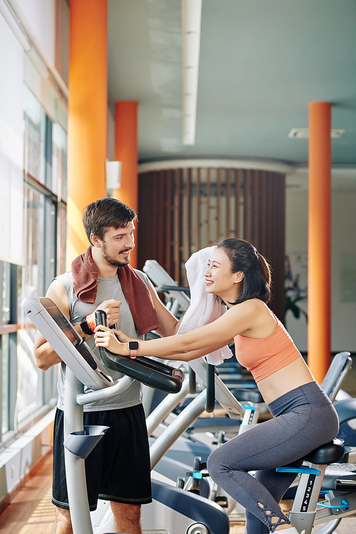 Fitness coach supporting client riding on spinning bike wiping sweat from her forehead with fluffy towel