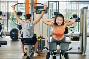 Cheerful young pretty Asian woman doing hypertension in gym when her boyfriend doing exercise at lat pulldown machine