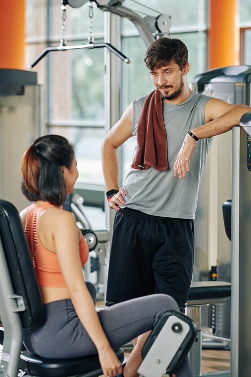 Handsome young coach talking to young woman doing hips exercise in abductor machine in gym