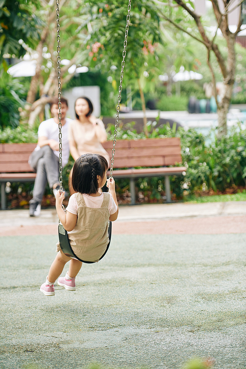 Little girl swinging when her parents resting on bench in park