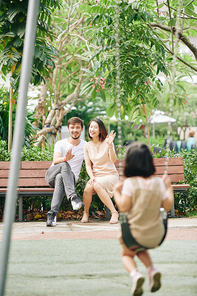 Happy multi-ethnic couple sitting on bench and waving to their little daughter sitting on swing