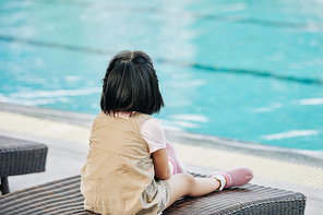 Pensive little girl sitting on chaise-lounge and looking at turquoise water in swimming pool
