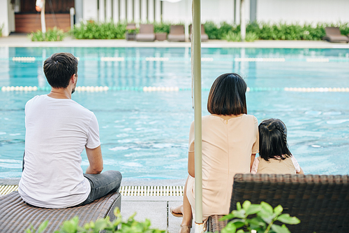 Young family resting by swimming pool and looking at clean turquoise water, view from the back