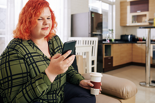 Plus size young woman with bright orange hair sitting on sofa at home, drinking coffee and checking messages on smartphone