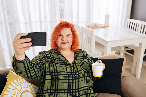 Smiling young woman in plaid shirt sitting on couch with cup of coffee and taking selfie