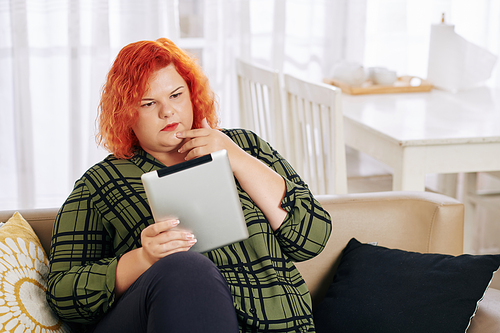 Pensive young pluz size woman sitting on sofa and reading article on tablet computer