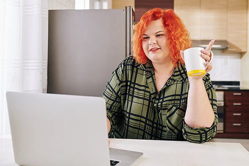 Smiling young plus size woman drinking morning coffee and reading news article on laptop screen