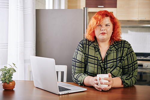 Portrait of pretty plus size young woman sitting at kitchen table with opened laptop, drinking coffee and looking at camera