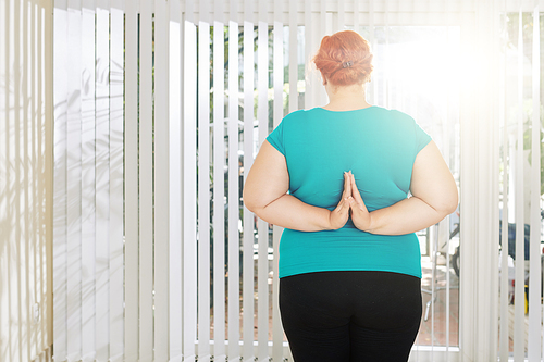 Standing plus size woman doing reverse prayer pose to strengthen her arms, view from the back