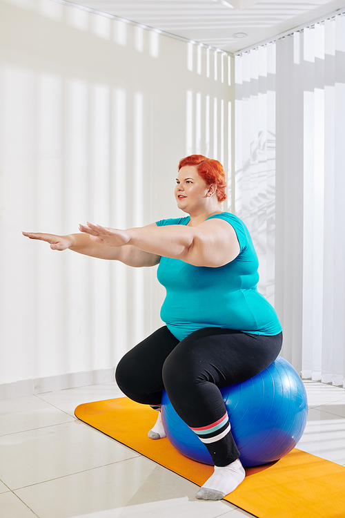 Young positive plus size woman holding arms in front of her when balancing on fitness ball