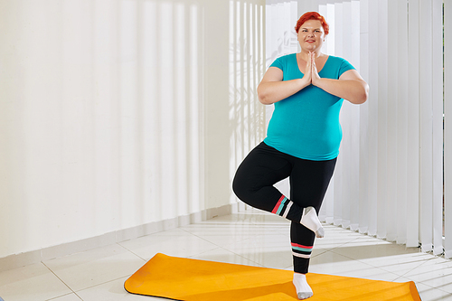 Happy pretty young plus size woman standing in tree pose on yoga mat and looking at camera