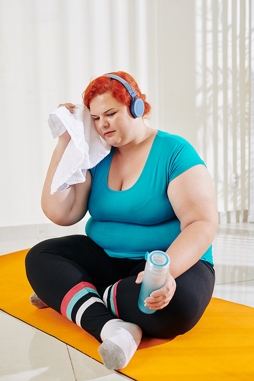 Tired plus size young woman sitting on yoga mat with bottle of water and wiping sweat from her forehead