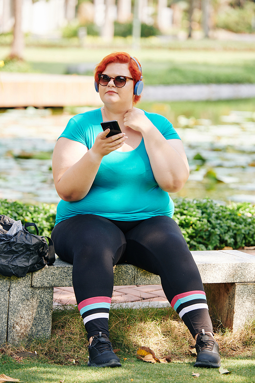 Plus size young woman resting on bench in park and choosing music on smartphone after jogging in the morning