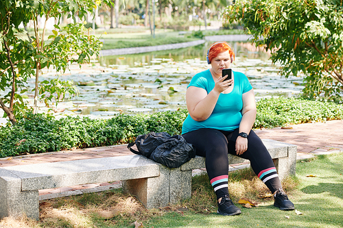 Plus size young woman sitting on bench in park and listening to music in her smartphone