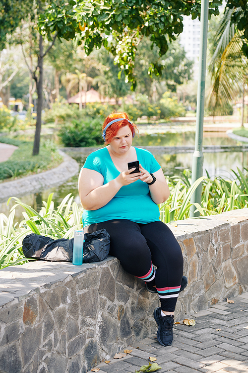 Young plus size woman resting on stone wall in park and checking messages and notifications in her smartphone