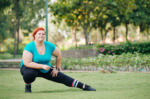 Plus size young woman stretching legs and warming up after jogging in park