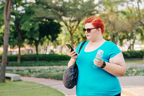 Yuong plus size woman walking in park with bottle of fresh water and checking messages and notifications in smartphone