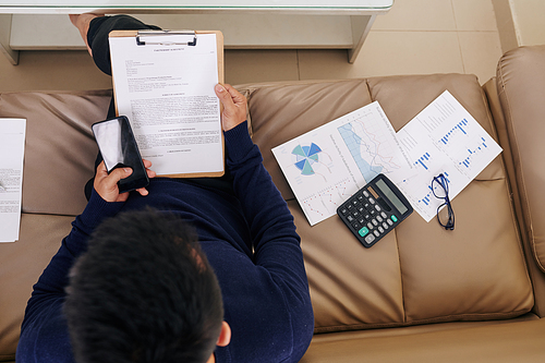 Businessman sitting on sofa with financial charts and reading partnership agreement, view from above