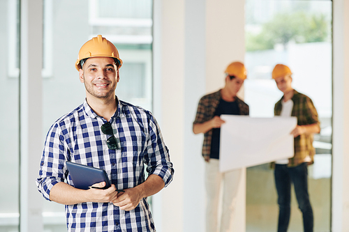 Medium portrait of professional engineer wearing casual shirt and hardhat holding tablet looking at camera, his colleagues looking at building plan