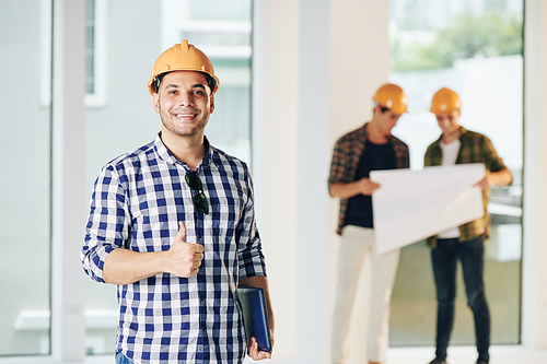 Medium portrait of construction engineer wearing casual shirt and hardhat looking at camera showing thumb up, his colleagues looking at building plan