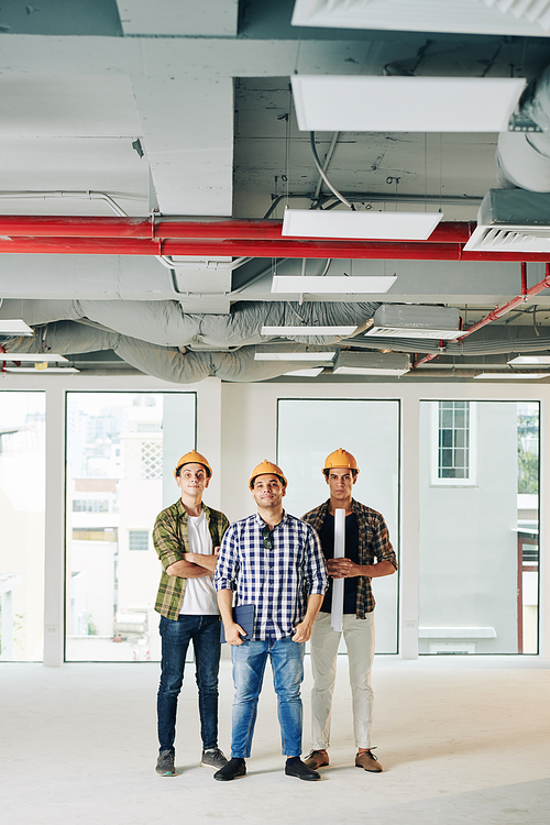 Vertical long shot of three professional engineers  wearing checked shirts and hardhats standing together inside new building space