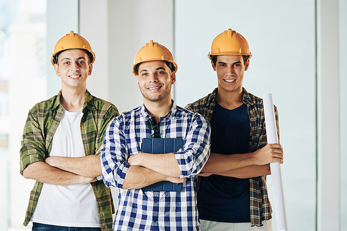 Horizontal medium shot of three professional engineers wearing checked shirts and hardhats standing together with arms crossed looking at camera smiling