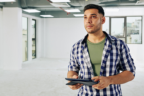 Horizontal waist up portrait of young adult man holding digital computer looking around his future office room
