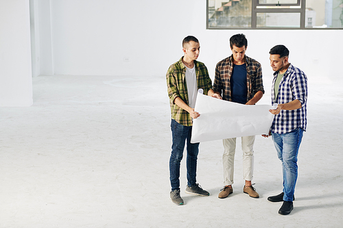 Group of three young adult engineers standing in empty spacious room looking at building plan paper, horizontal high angle long shot, copy space