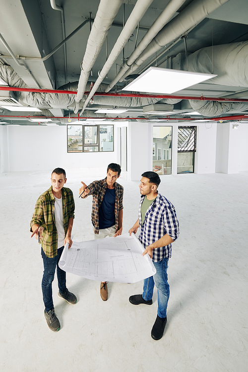 Group of professional engineers standing in empty spacious room holding building plan paper doing brainstorming, vertical high angle long shot, copy space