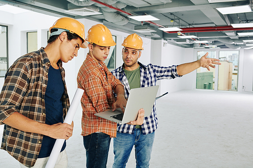 Three modern construction workers wearing hardhats searching additional information for current building project using laptop, copy space