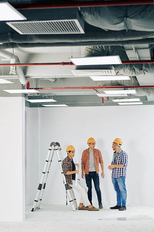 Vertical long shot of three professional construction engineers gathered together in unfinished room talking to each other, copy space
