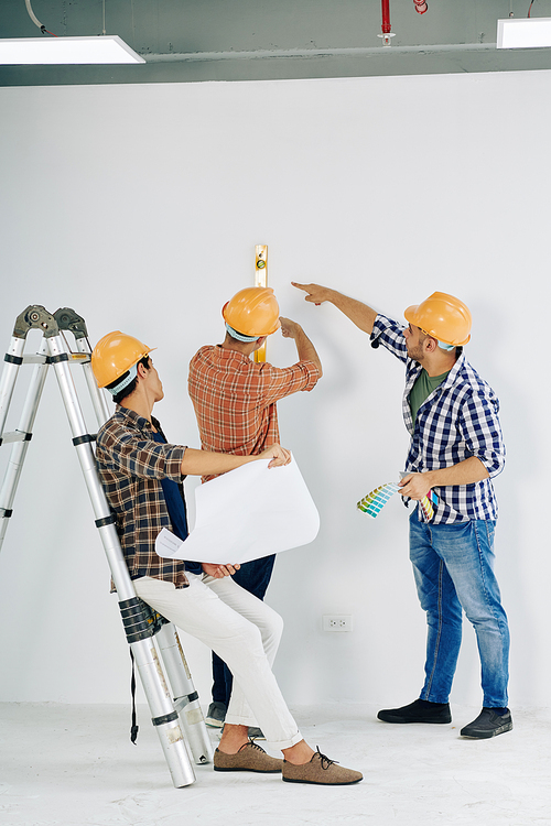 Modern manual workers wearing yellow hardhats checking unfinished room wall level, vertical shot
