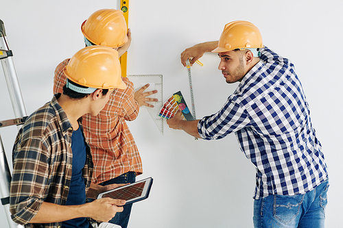 Group of three modern manual workers wearing checked shirts and yellow hardhats taking measurements for further work