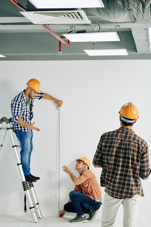 Vertical shot of professional construction worker standing on stepladder holding tape rule measuring wall length, his colleagues helping him, copy space