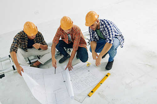 Horizontal high angle shot of modern construction workers sitting on hunkers looking into building plan papers, copy space