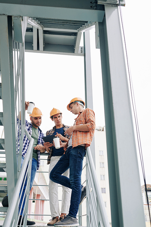 Vertical low angle shot of three modern professional construction engineers standing relaxed upstairs chatting during coffee break, copy space