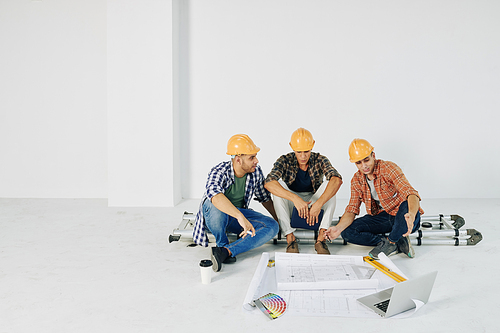 Middle Eastern men wearing casual clothes and hardhats sitting together in empty room with papers and laptop working on building plan
