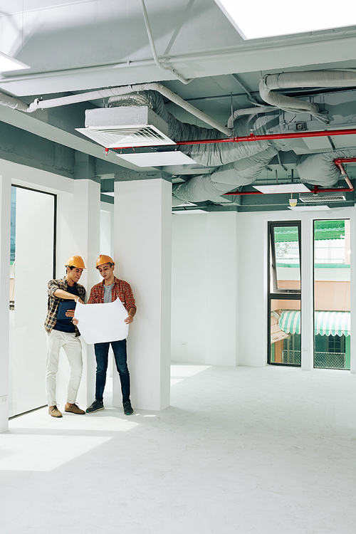 Two modern construction engineers standing together in unfinished room looking into building plan paper, copy space