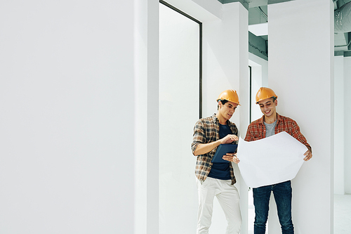 Two professional construction engineers standing together in unfinished room working with building plan paper, copy space