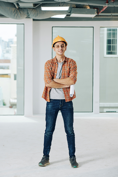 Full body shot of confident construction worker wearing casual outfit and protective hardhat standing with arms crossed looking at camera