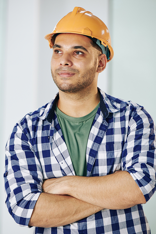 Waist up shot of young adult construction engineer wearing checked shirt and protective hardhat standing with arms crossed looking away