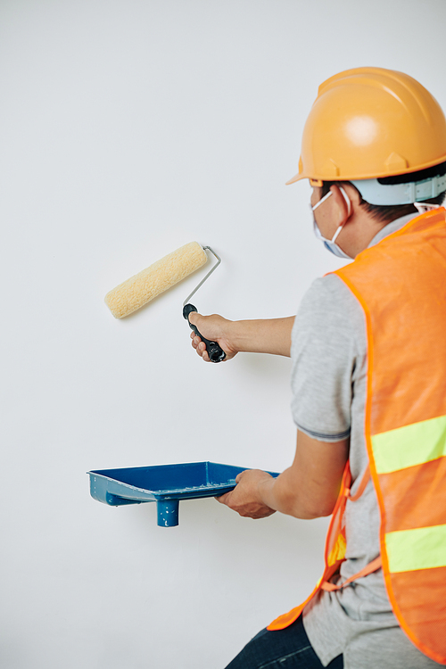 Worker in hardhat and protective mask using plastic tray and fluffy roll when painting walls white