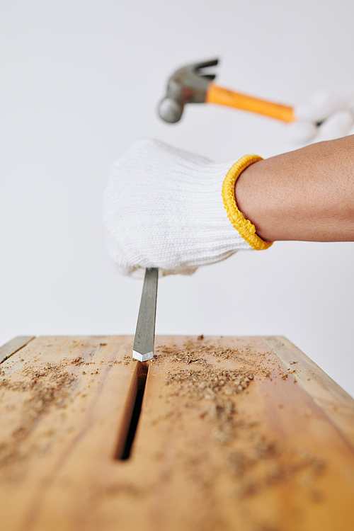 Carpenter in textile gloves using chisel and hammer when making hole in wooden bench