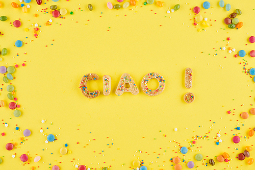 Ciao inscription made of sweet homemade cookies on yellow background with chocolate candies and colorful sprinkles