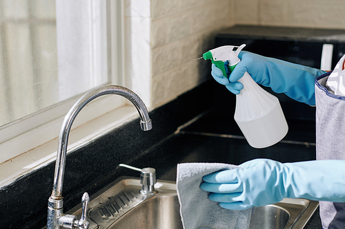 Housewife spraying kitchen tap with cleaning detergent and wiping off dirt and stains